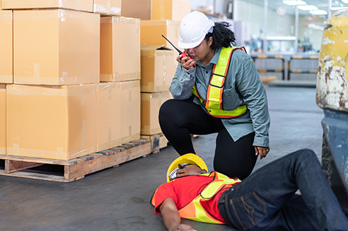 Warehouse worker lying down on floor after having accident from working in the factory. Female manager using walkie talkie calling colleagues to help injured man.